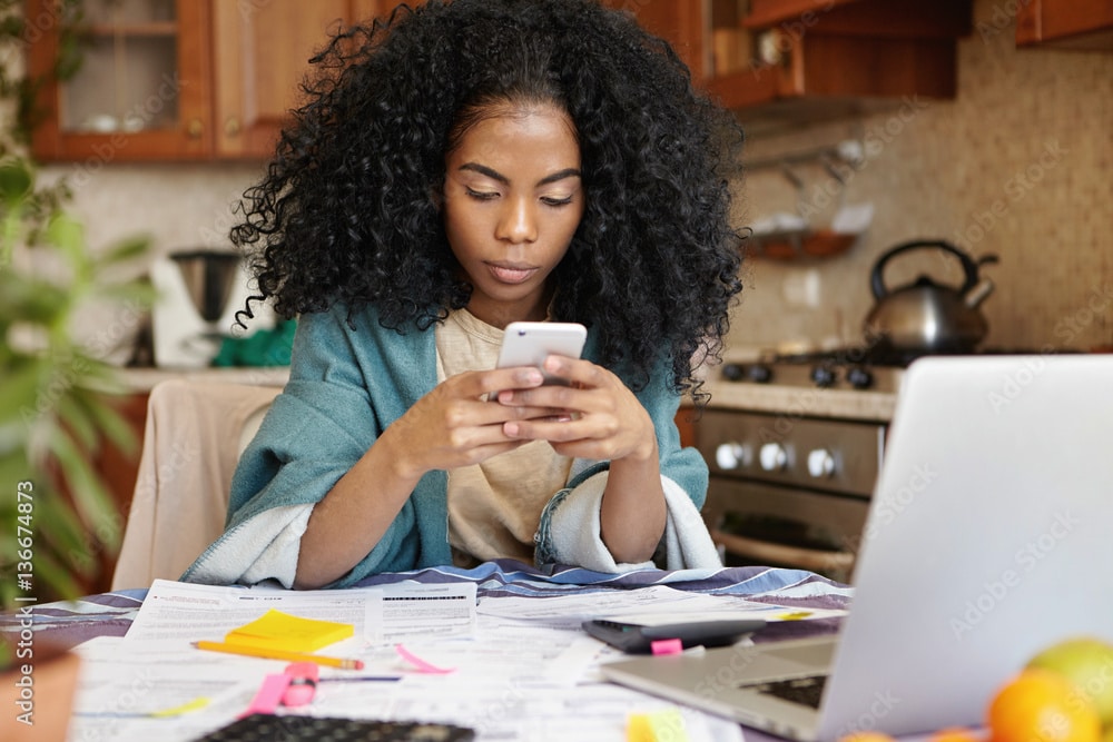 Indoor shot of unhappy young lady using mobile phone in front of laptop and analyzing home finances and credit card bills.