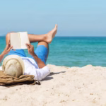 A handsome man relaxing on a beach reading a book and thinking about what he would do with financial freedom.