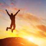 Happy man jumping for joy on the peak of the mountain representing a high ranking on the global happiness report.