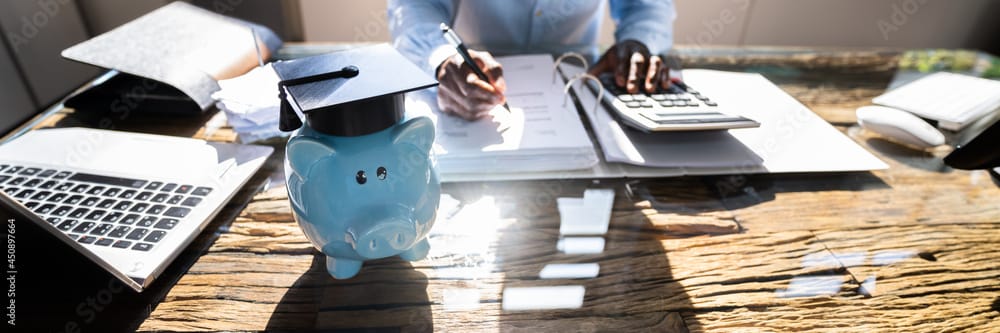 Picture of piggy bank on desk with person working on Student Loan options as learned on Think and Talk Money.