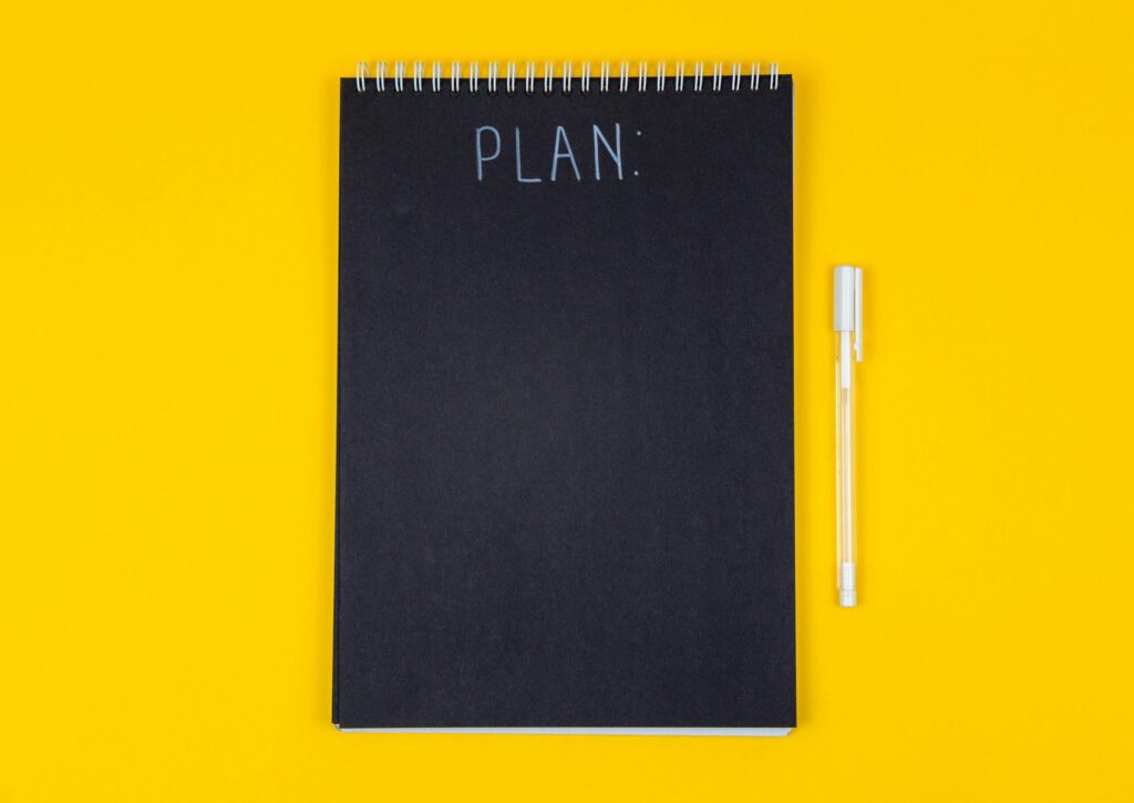 Wide view image of blank black spiral note pad and white marker with calligraphic inscription plan on yellow background meaning we all need a plan to keep our money.