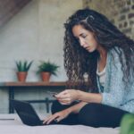 Young woman is making web payment holding bank card and working with laptop sitting on bed at home indicating that using cash back is not the best redemption option for your points.