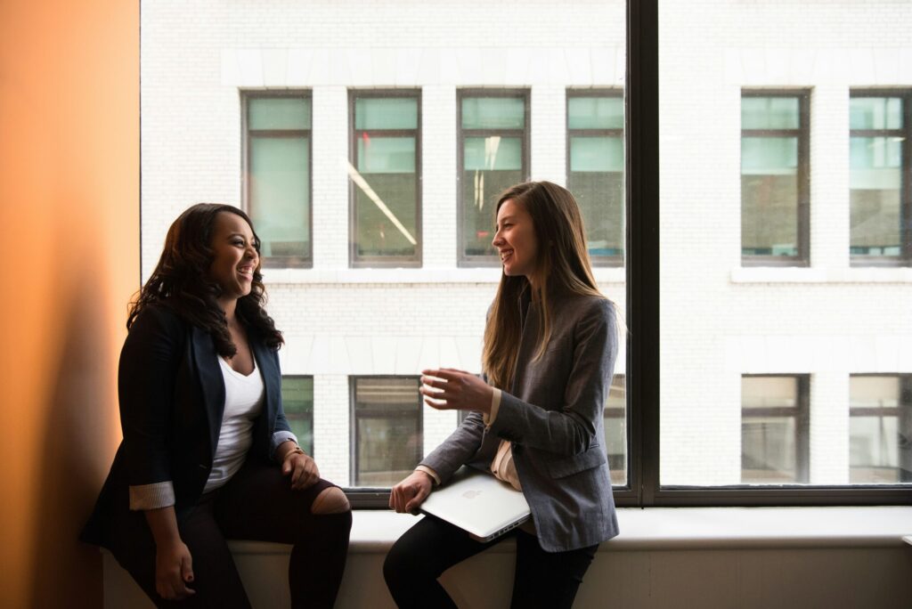 two women sitting by the window talking about investing when they really need to learn about the basics of personal finance.